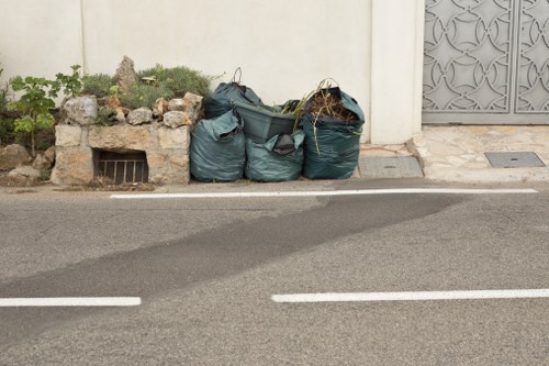 The image shows three large dark green refuse bags filled with rubbish, positioned on the pavement close to the edge of a road in an urban area. The bags are made of thick plastic with a slightly glossy finish and are partially open, revealing some tangled materials inside. To the left of the bags, there is a small garden bed bordered by a rough stone wall, containing some green plants and dried twigs. Behind the bags, a white wall with a small, square metal grate near the ground is visible. On the right side of the image, there is a decorative grey metal gate with intricate patterns situated within a white wall, indicating a residential or commercial property boundary. The setting appears to be daytime, with natural lighting illuminating the scene. This arrangement of rubbish bags on the pavement aligns with waste removal services such as those offered by Office Clearance Knightsbridge, serving local areas including Knightsbridge and surrounding London districts.