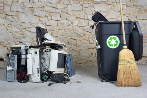 A pile of electronic waste and broken computer equipment, including discarded monitors, keyboards, and desktop towers with visible cables and plastic casings, stacked haphazardly on a paved surface in front of a textured stone wall. To the right, a black recycling bin with a green recycling symbol on its front is placed next to a wooden-handled broom leaning against it. The environment suggests a courtyard or driveway area where a rubbish removal service, such as Office Clearance Knightsbridge, might collect large electronic items and general waste for disposal. The scene is well-lit with natural daylight, emphasizing the contrast between the metallic and plastic textures of the electronic items and the rough stone background, highlighting the importance of proper waste collection and recycling in the local area, including Knightsbridge.