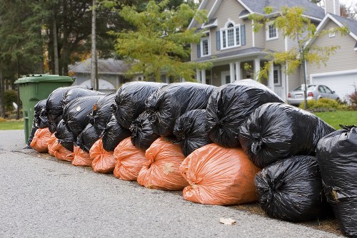 A row of tightly packed large black and orange plastic rubbish bags arranged along the edge of a residential street in an urban neighbourhood, likely for collection or disposal. The bags are resting on a paved asphalt surface, with the black ones positioned directly on the ground and the orange ones placed underneath, showing their bright, slightly glossy material. Behind the row, there are green wheelie bins with lids, positioned near some greenery and trees, suggesting a typical household waste collection outside a residential property. In the background, a light-colored, multi-storey house with a gabled roof, large windows, and a neatly maintained front garden is visible. The setting appears during daytime with natural light, and the street is lined with other similar homes, indicating a suburban area in or near Knightsbridge. This scene illustrates typical rubbish removal efforts by companies like Office Clearance Knightsbridge, serving local property owners or businesses in the area.