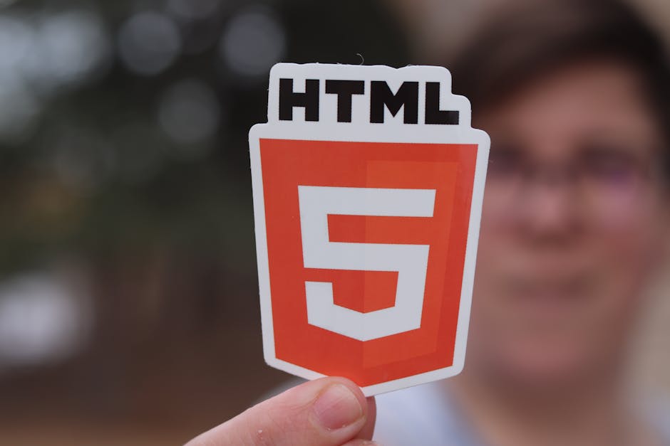 A person holding a small rectangular sticker featuring the HTML5 logo, which consists of a white number '5' inside an orange shield shape with a white border, and the word 'HTML' in bold black letters