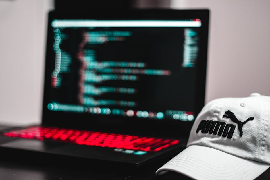 A close-up view of a person's hand typing on a silver laptop keyboard placed on a wooden desk. The laptop screen displays lines of code with syntax highlighting, indicating programming or web developm